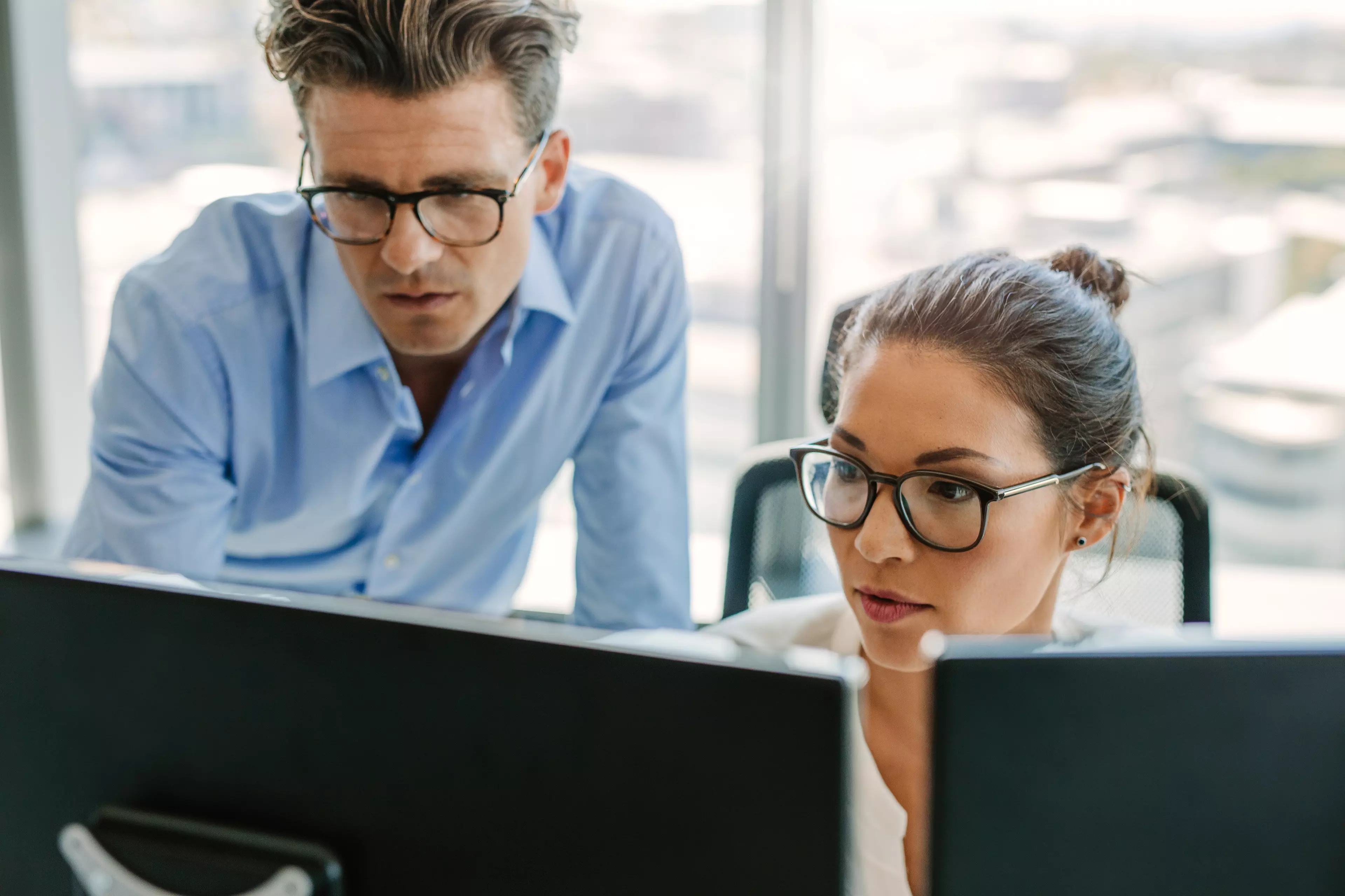 Woman in glasses, sitting behind two computer screens, showing something to a man next to her.