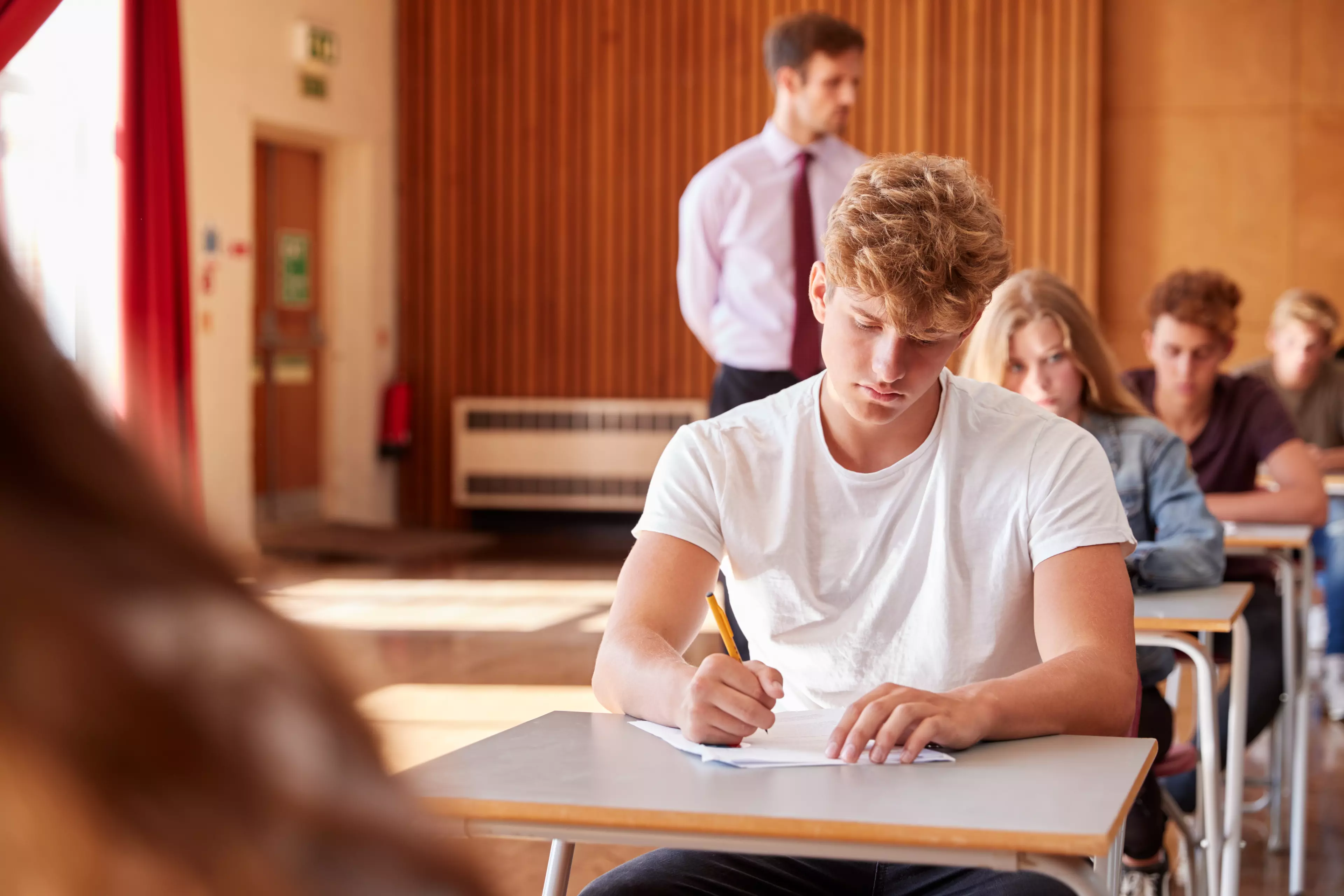  Young adult man sitting an exam in a room with other respondents, supervised by a proctor.