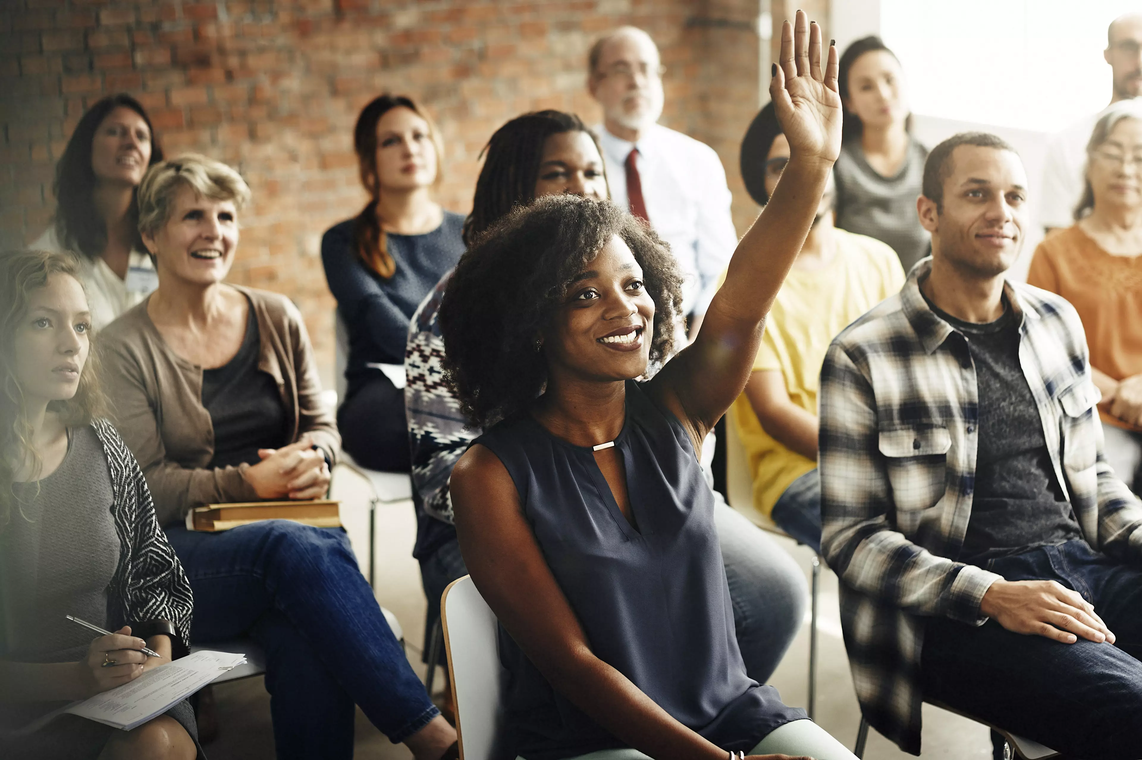 A group of smiling people during a meeting, with a young lady raising her hand to ask a question.