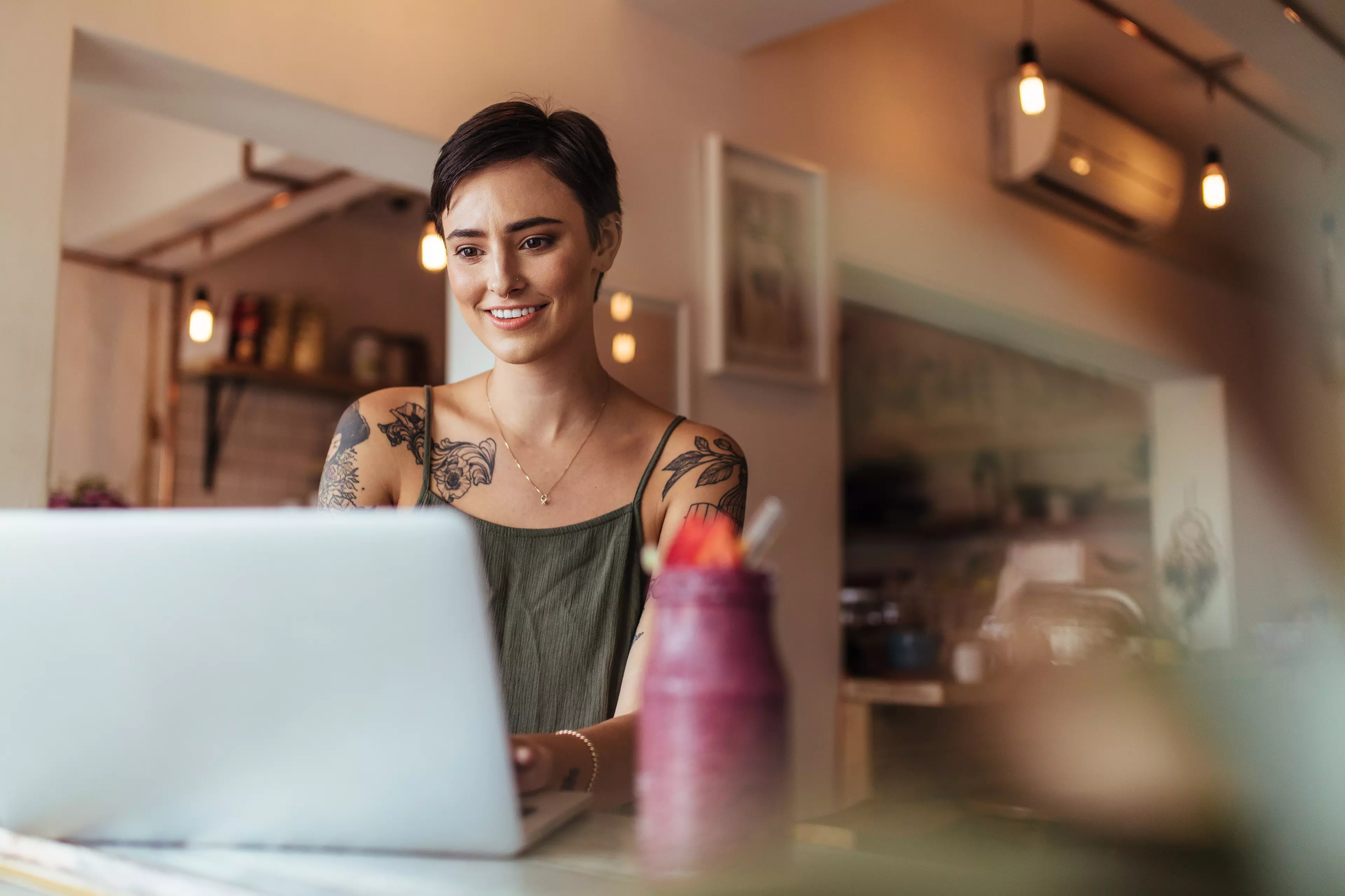  Short-haired, smiling woman with tattoos sitting at a desk, typing on her laptop.