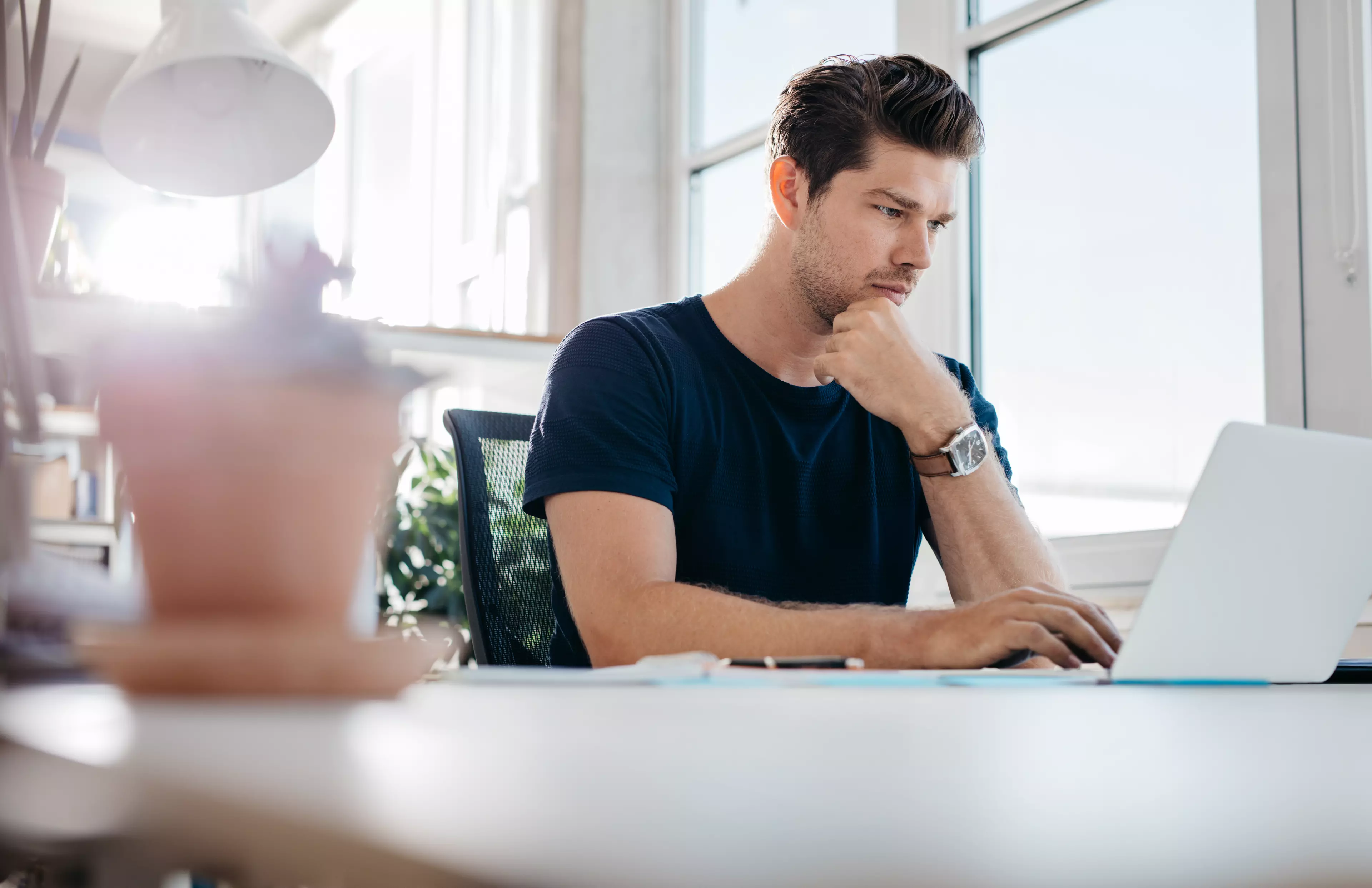  Young man sitting behind a table, looking at his laptop screen.