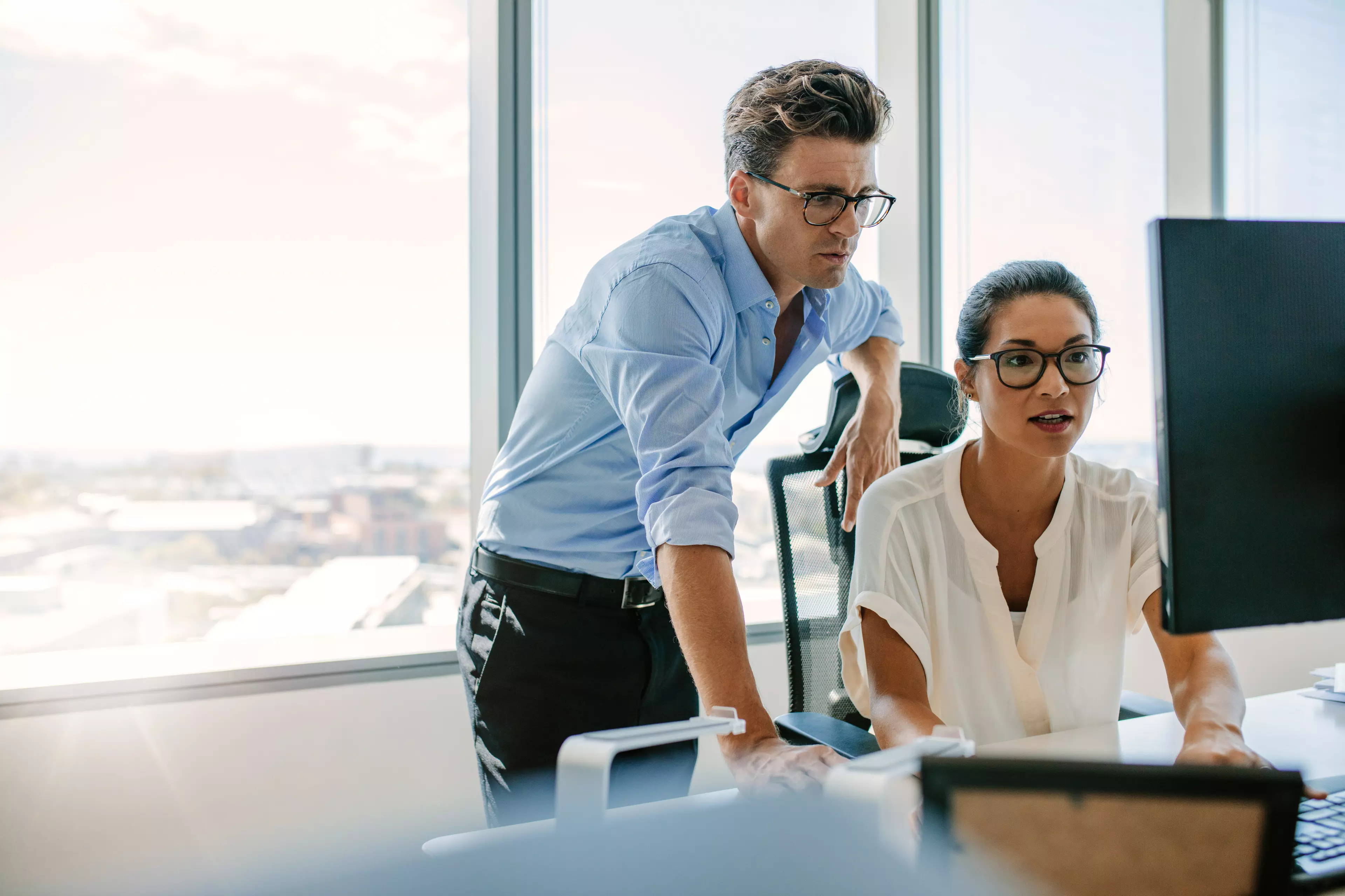  A sitting woman and a standing man in an office, looking at a computer screen.