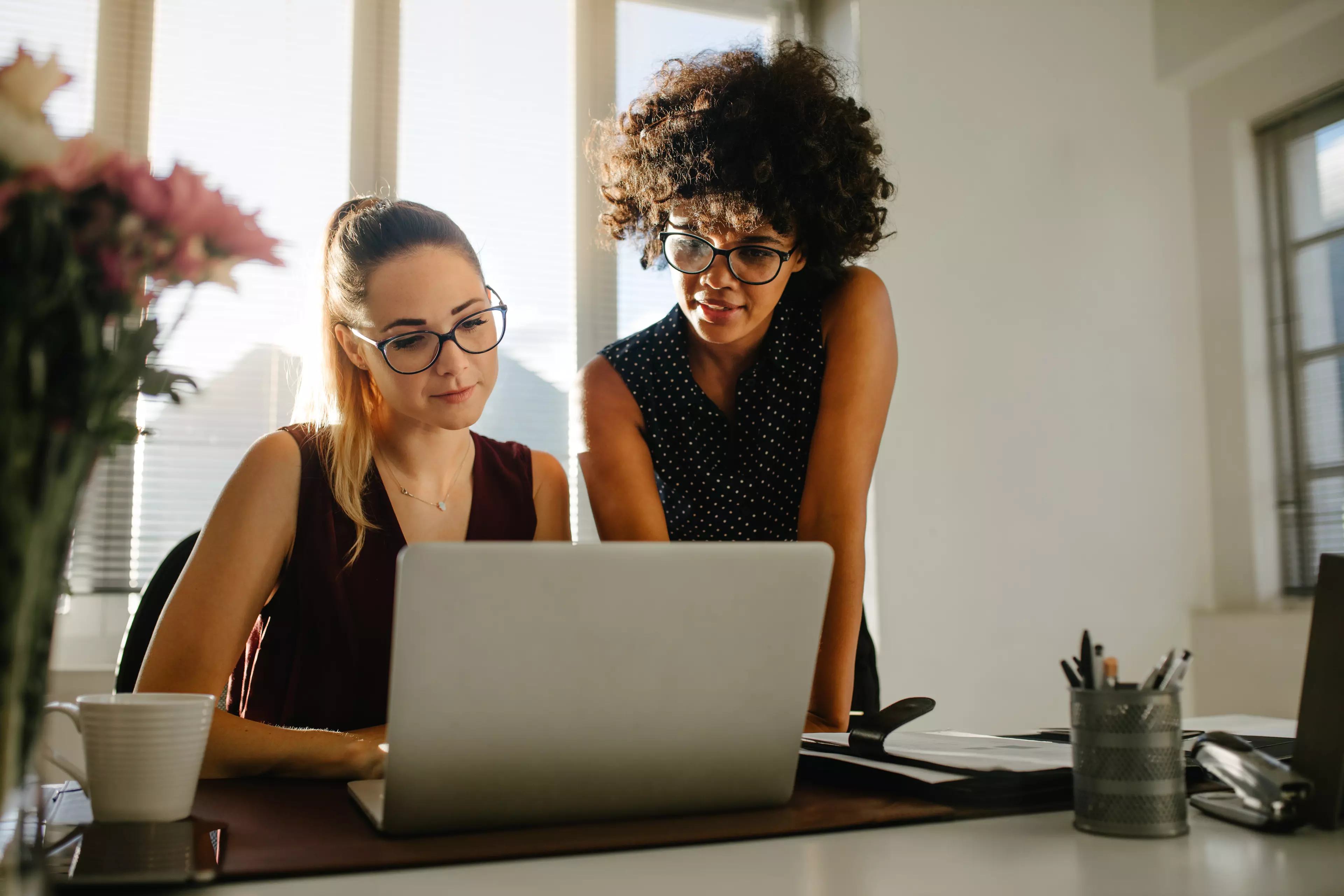 Two young women, one sitting and one standing discussing an image on a laptop.